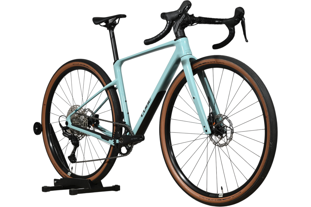 Velo gravel Loop Sports Cube Nuroad C:62 ONE 2025 bleu clair avec pneus a flancs marron sur support en bois